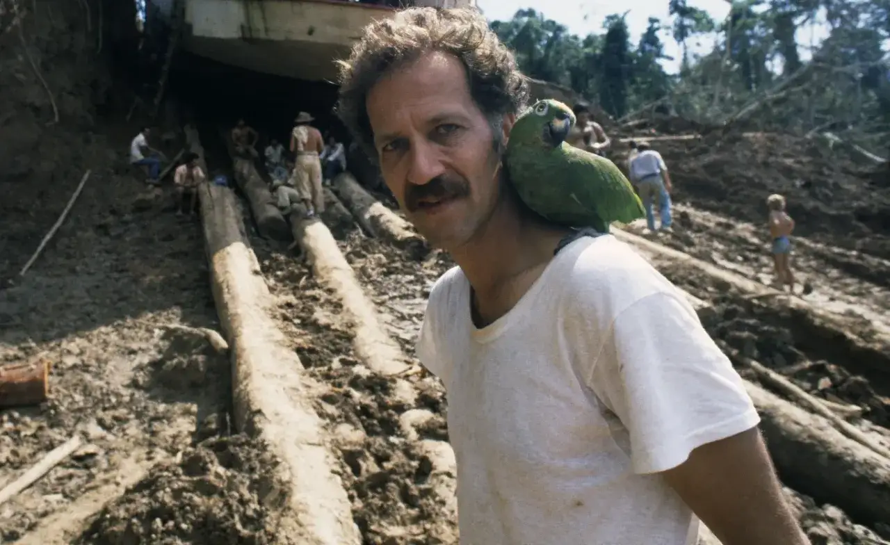 Werner Herzog z papugą na ramieniu, w tle budowa w dżungli, jak w jego filmach.