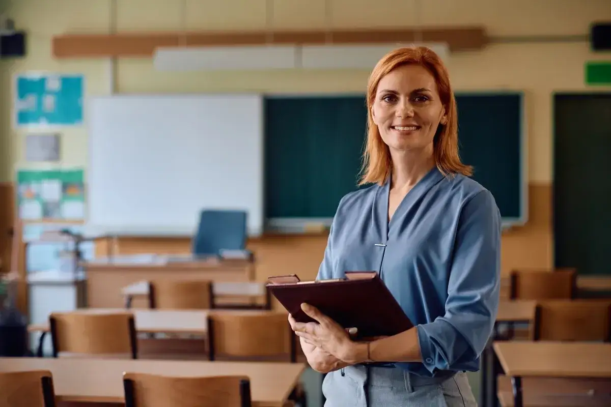 Uśmiechnięta nauczycielka z rudymi włosami trzyma teczkę. W tle klasa z ławkami i tablicą. Posiada legitymację nauczyciela.