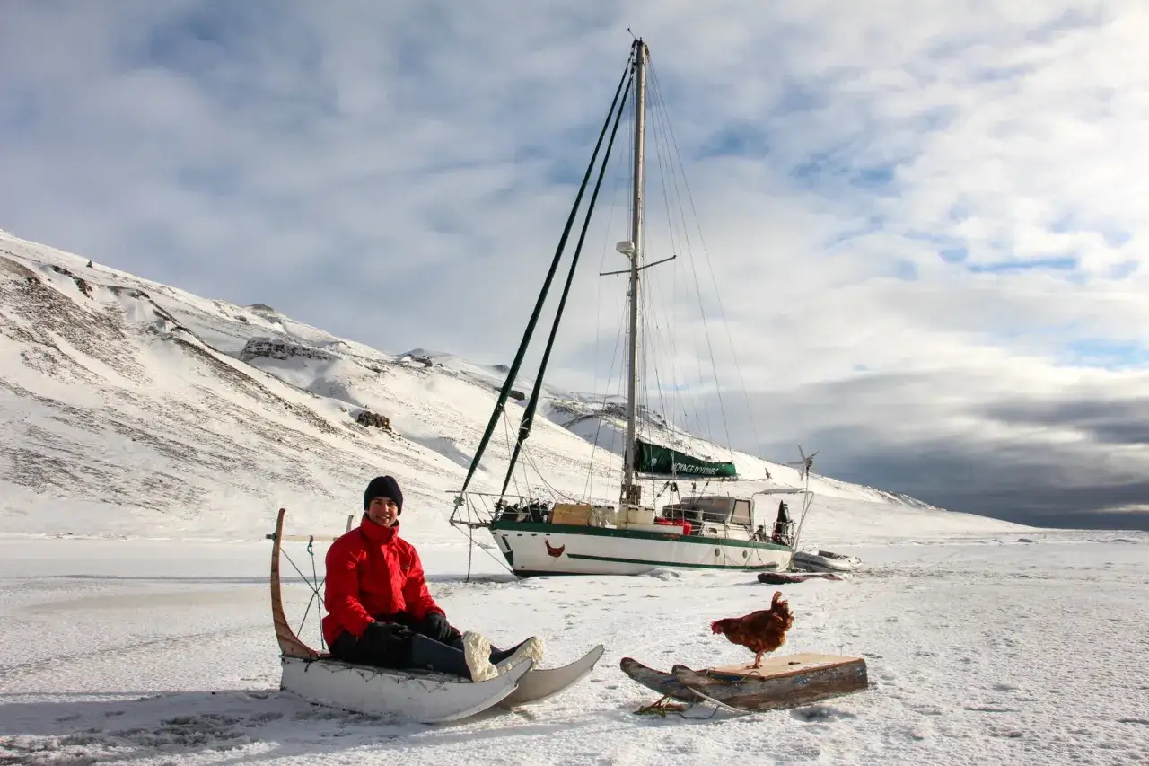 Guirec Soudée sitzt in einem Schlitten vor seinem Segelboot in einer verschneiten Landschaft. Eine Henne steht auf einem Holzbrett daneben.