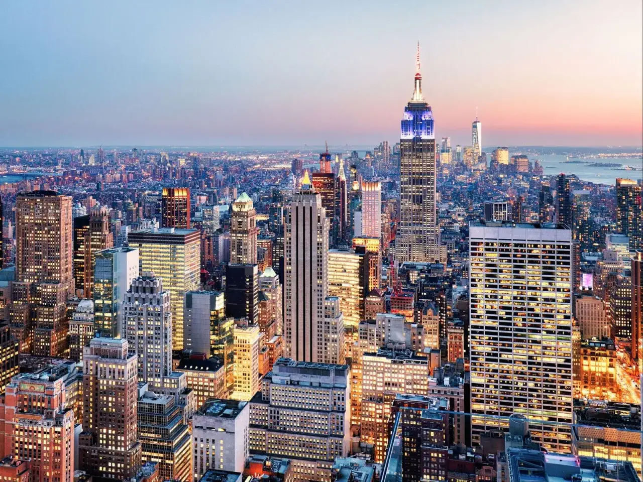 Widok na rozświetlony Nowy Jork o zmierzchu, z Empire State Building w centrum. Zastanawiasz się, ile kosztuje lot do USA?