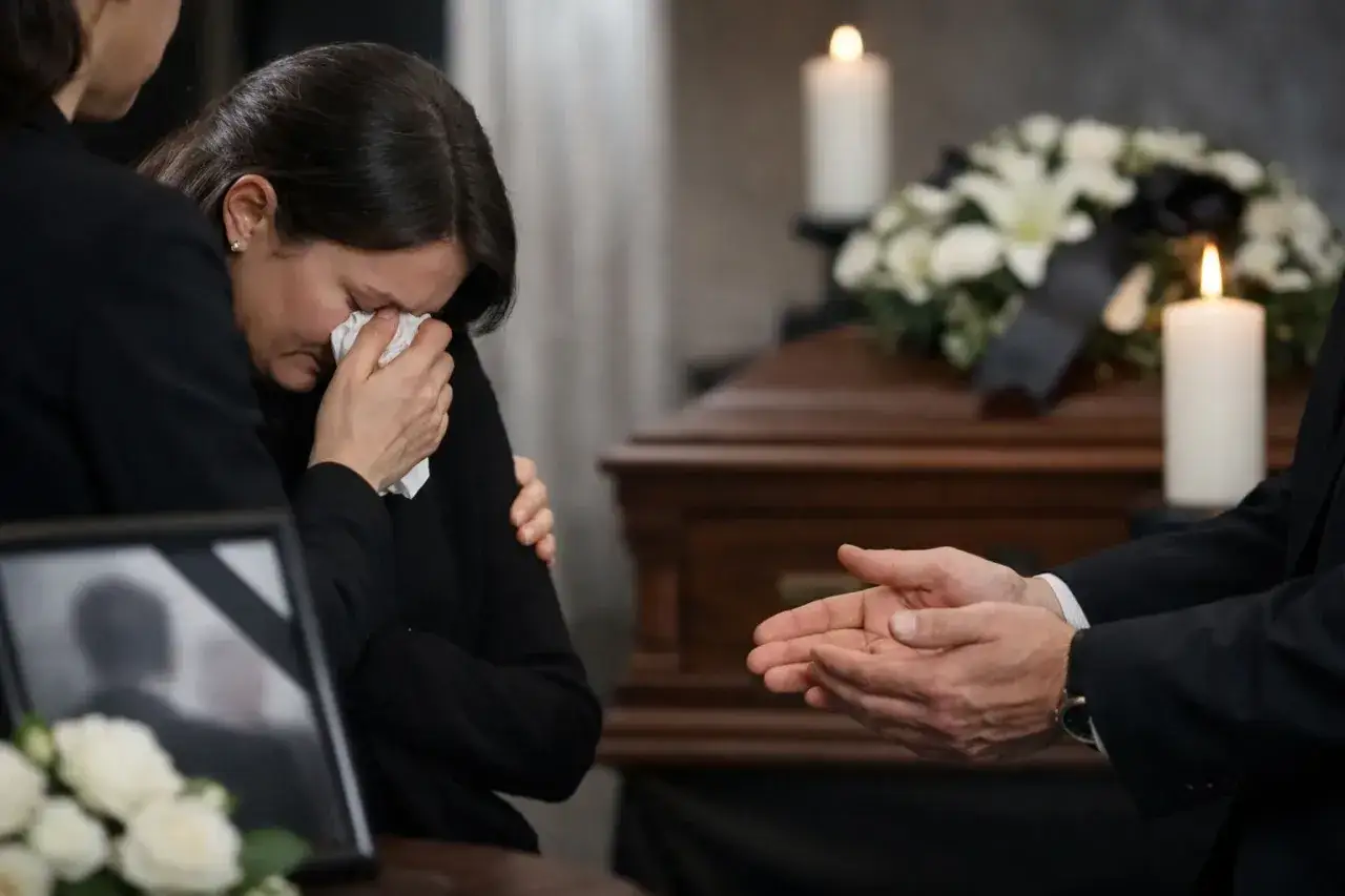 Smutna kobieta ociera łzy chusteczką, wspierana przez bliską osobę. W tle trumna, kwiaty i świece. Słowa pocieszenia na pogrzebie rodzinie.