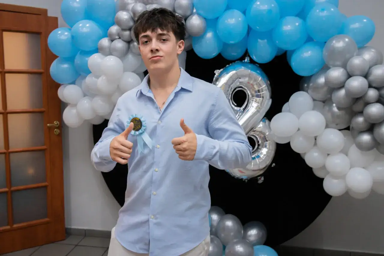 Chłopak w błękitnej koszuli z rozetką "18th Birthday" i balonami.
