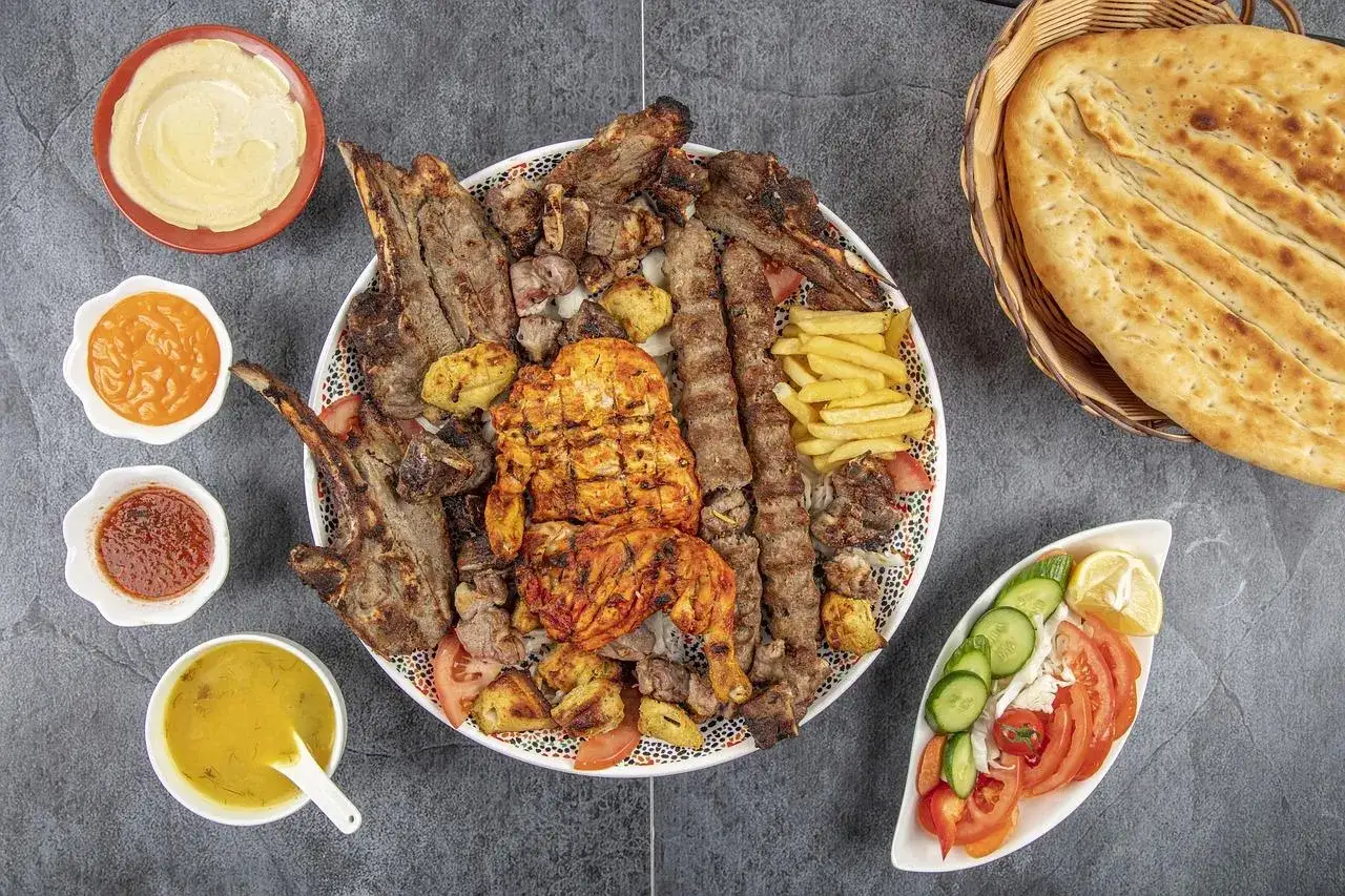 Talerz grillowanej baraniny, kurczaka, frytek, sałatki i sosów, obok chleba.