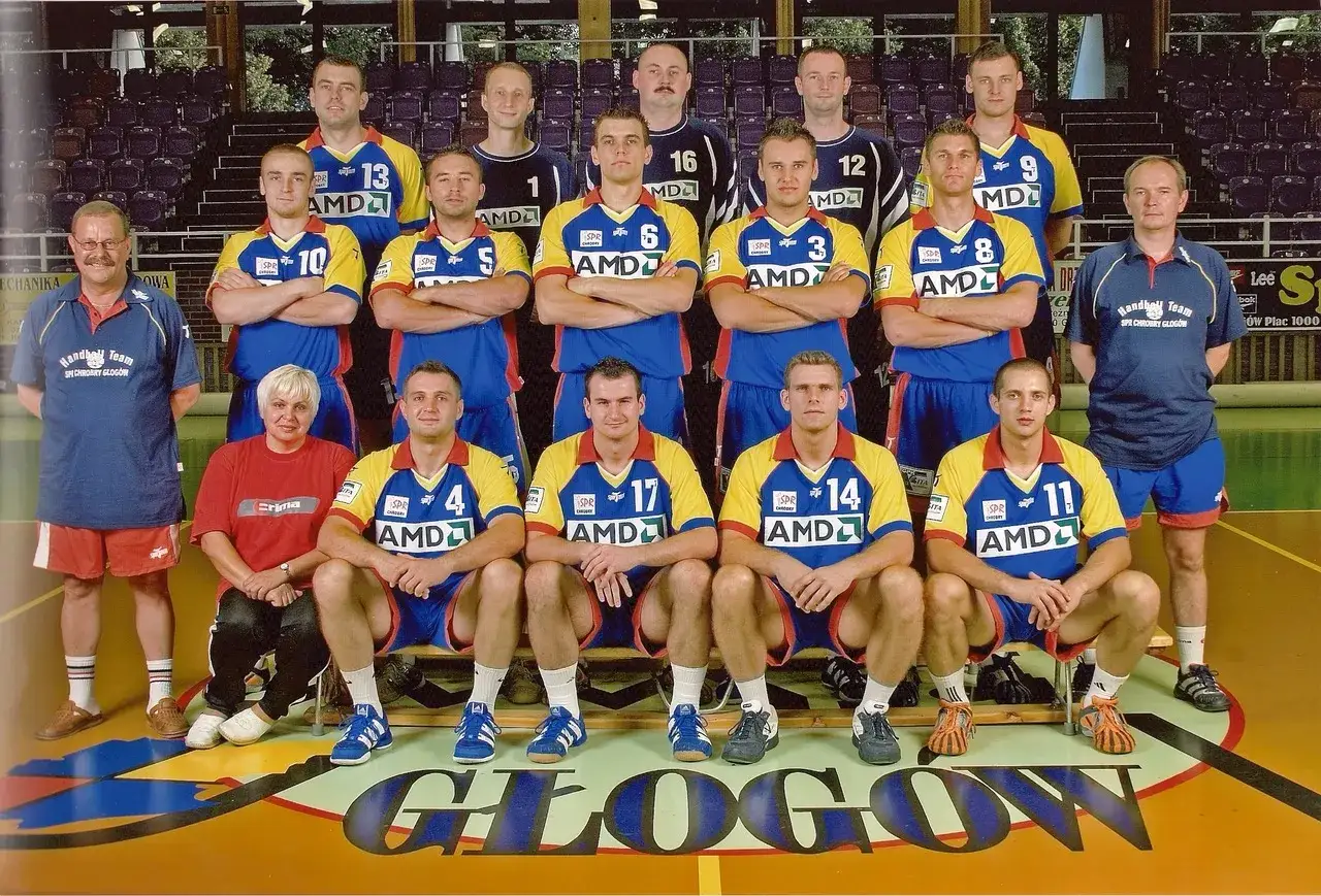 Drużyna SPR Chrobry Głogów pozuje do zdjęcia. Zawodnicy w żółto-niebieskich strojach sportowych, trenerzy i jedna kobieta.