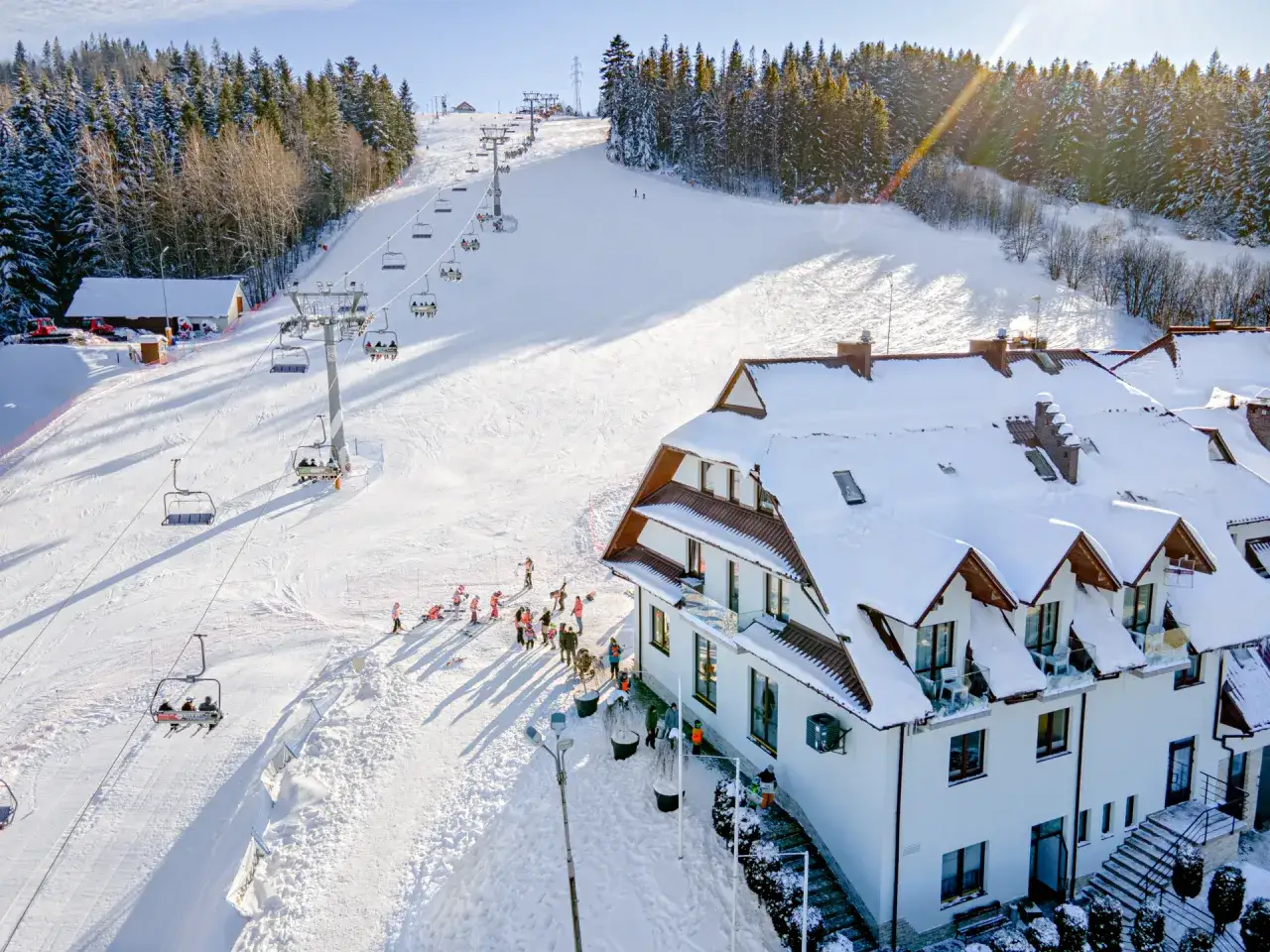Hotel ze stokiem narciarskim dla dzieci. Kolejka krzesełkowa wwozi narciarzy na ośnieżony stok, a obok hotelu bawią się dzieci.