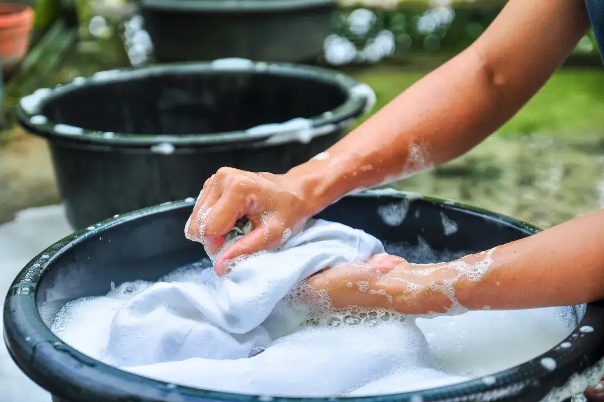 Manos lavando ropa blanca a mano en una tina con espuma, un método tradicional que explica qué significa lavado a mano en lavadora.