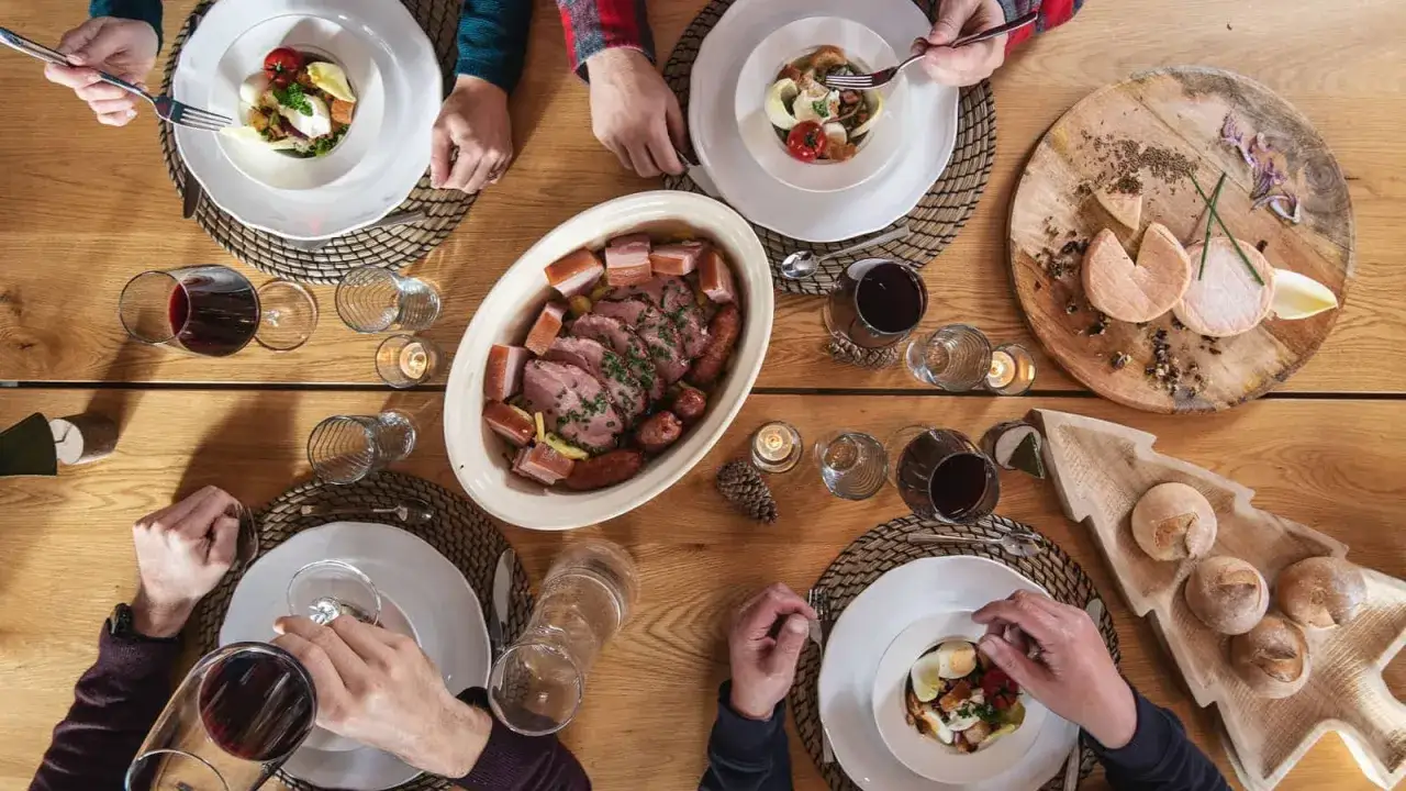 Un repas du terroir avec viande, saucisses, fromages et pain sur une table en bois. Des mains servent et dégustent.