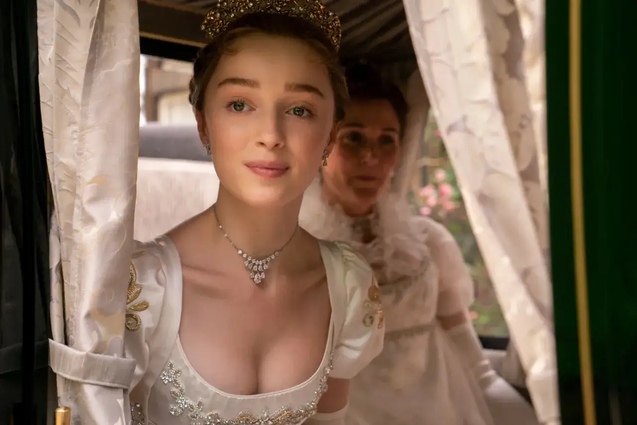 Phoebe Dynevor w stroju z epoki, z tiarą i naszyjnikiem, w karocy. Jej filmy, seriale i programy to często historie kostiumowe.