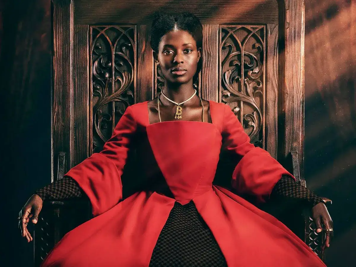 Jodie Turner-Smith, con su mirada regia, se sienta en un trono de madera tallada, vestida con un opulento traje rojo.