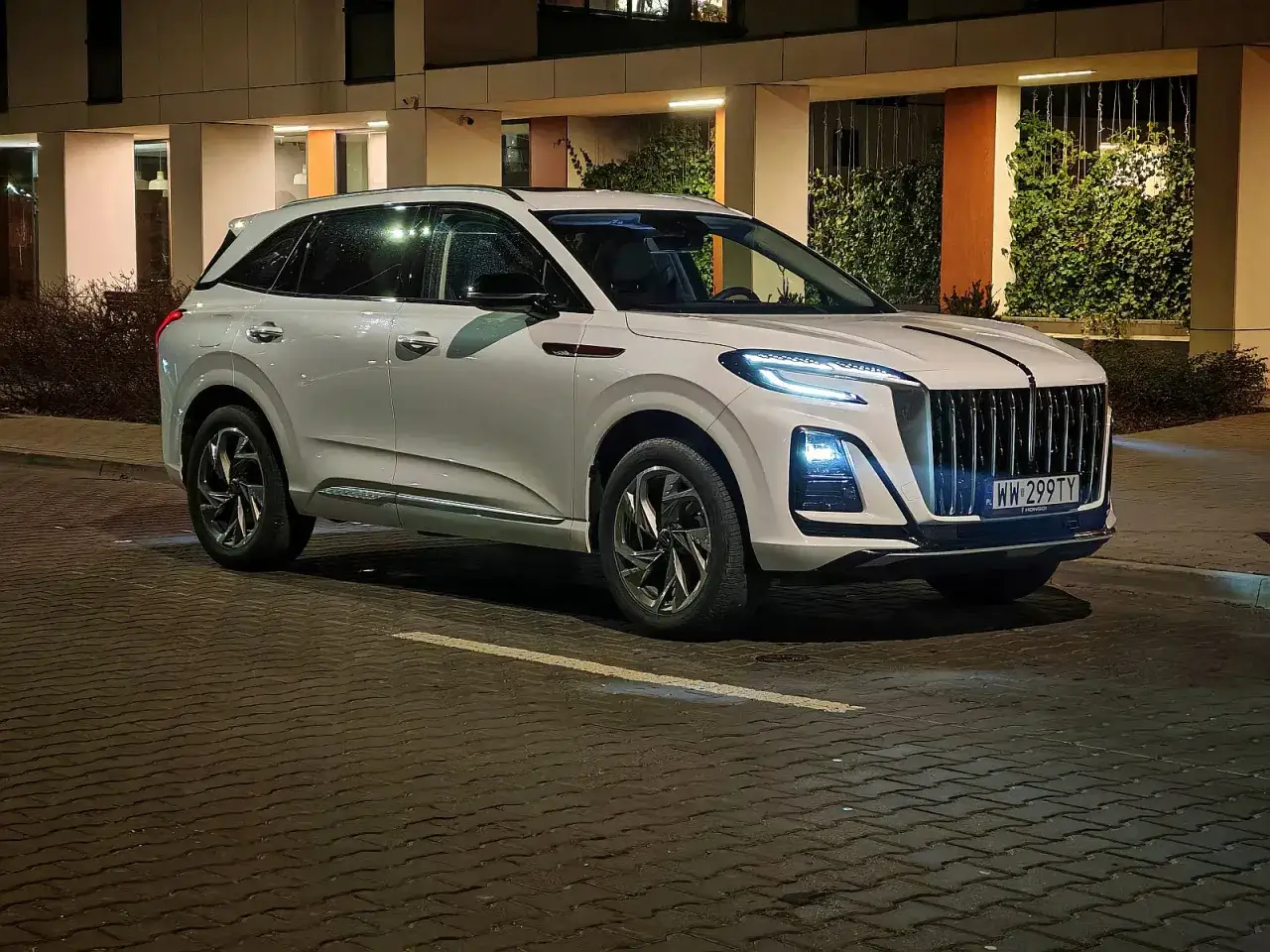 Biały Hongqi HS3 z charakterystycznym grillem i nowoczesnymi światłami stoi na mokrej, brukowanej ulicy nocą.