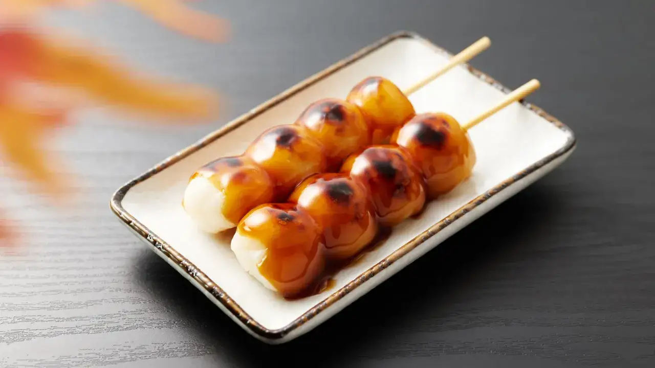 Brochettes de dango grillés, nappées de sauce sucrée, sur une assiette blanche.