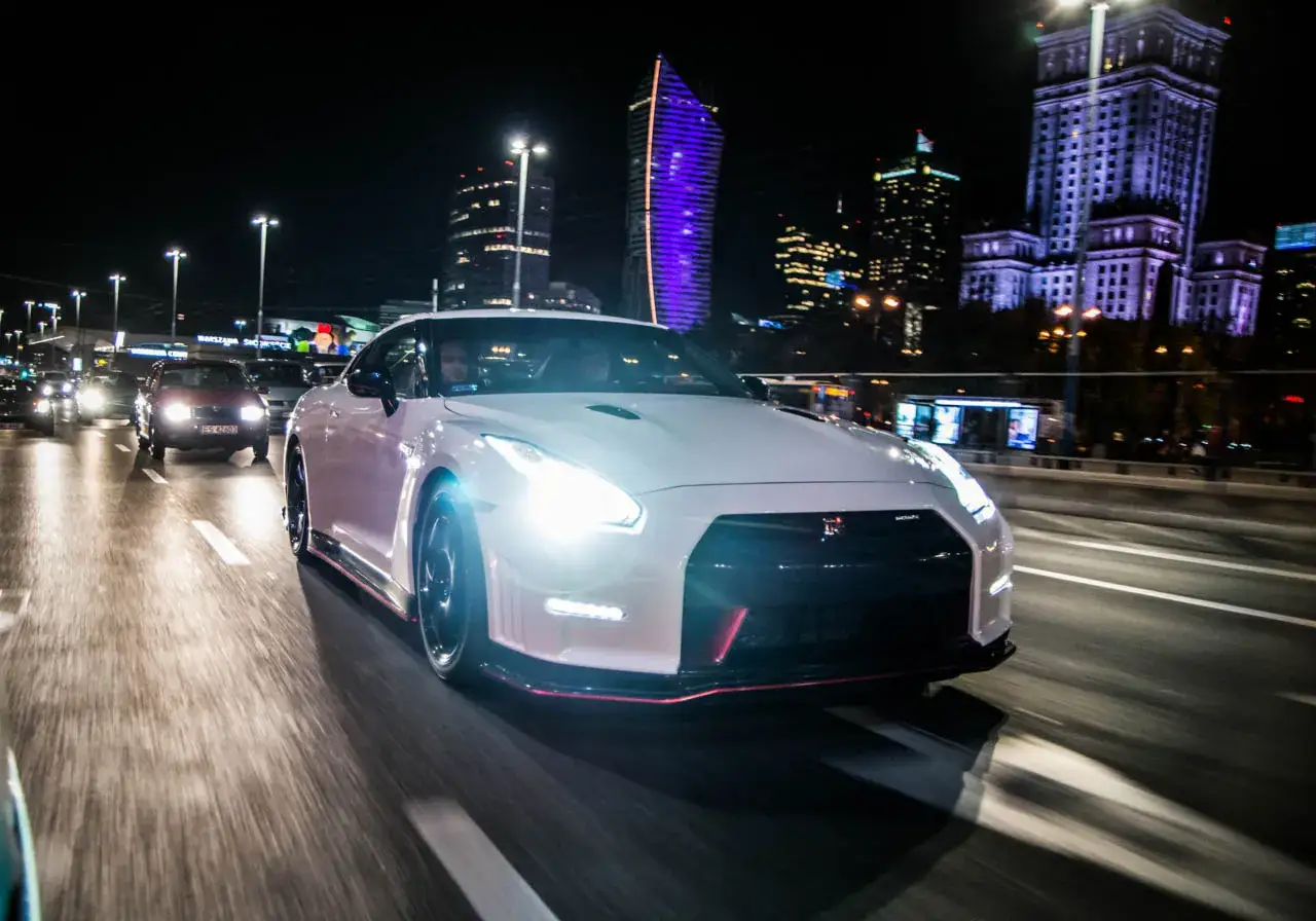Biały Nissan GT-R Nismo mknie nocą przez miasto, oświetlony reflektorami i neonami wieżowców.