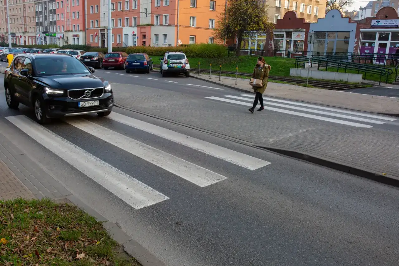 Kobieta z paczką przechodzi przez przejście dla pieszych. Samochód zatrzymał się, by ją przepuścić.