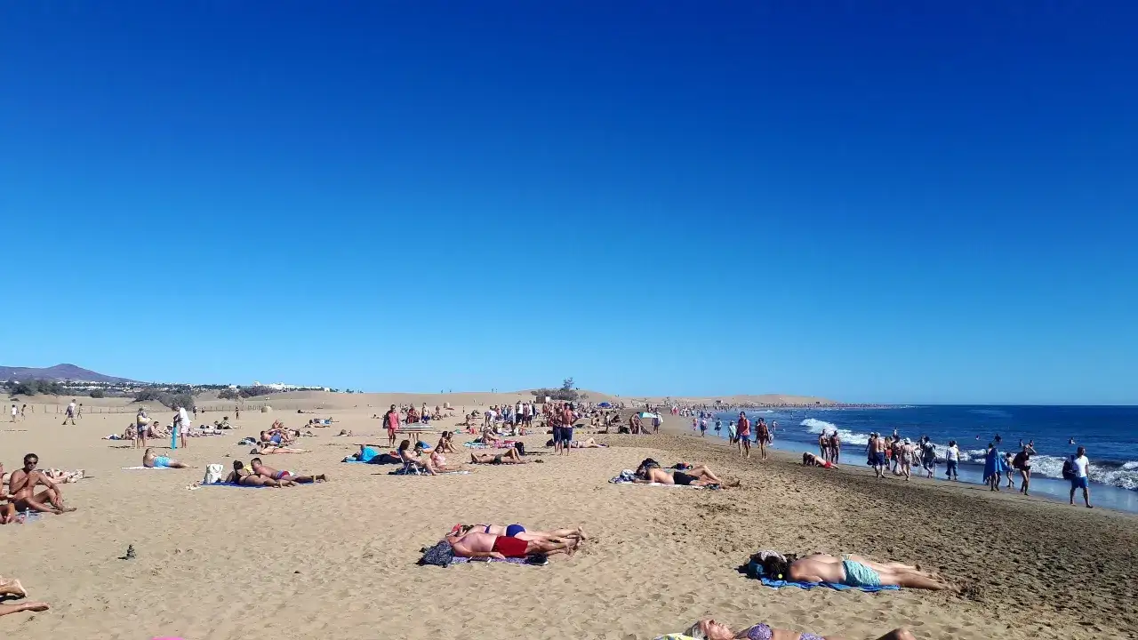 Tłum ludzi odpoczywa na słonecznej plaży. Woda jest spokojna, a niebo bezchmurne. Czy Gran Canaria jest bezpieczna? Tak, wygląda na to, że turyści czują się tu swobodnie.