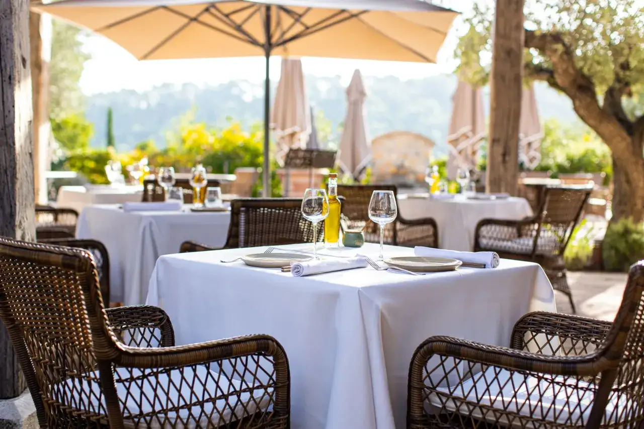 Tables dressées en terrasse, sous un parasol, invitant à savourer une cuisine du terroir authentique. Vins, verres et couverts attendent les convives.