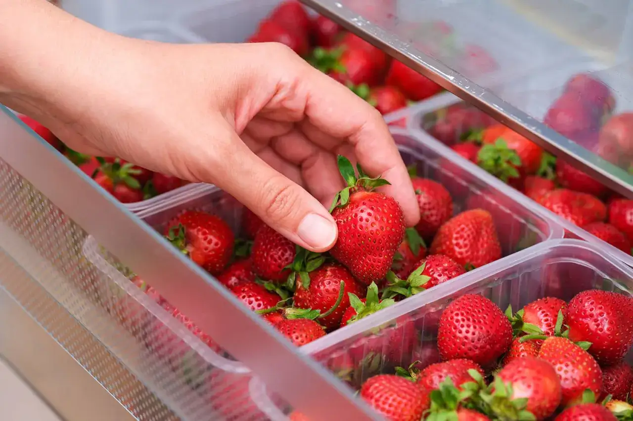 Hand wählt eine saftige Erdbeere aus einer Plastikschale. So lagert man Erdbeeren am besten, um sie frisch zu halten.