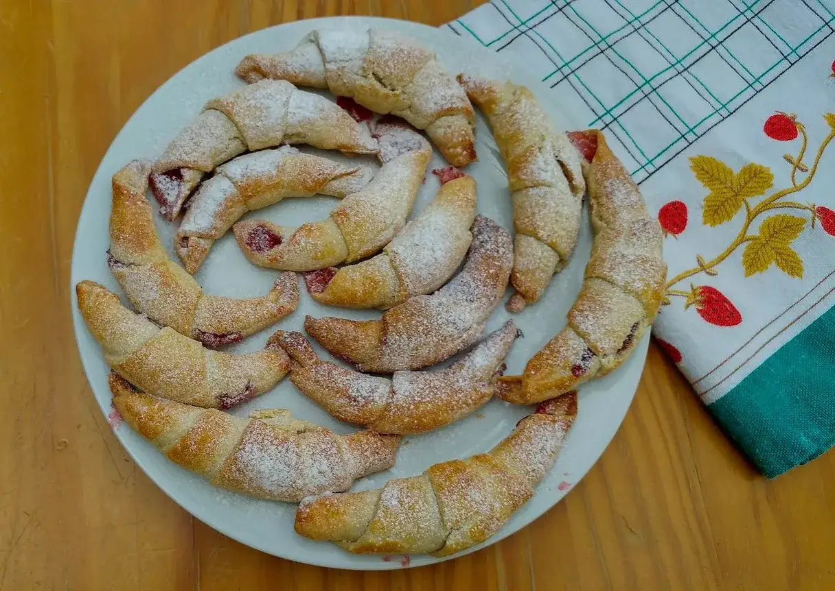 Kruche rogaliki z marmoladą, posypane cukrem pudrem, ułożone na talerzu.