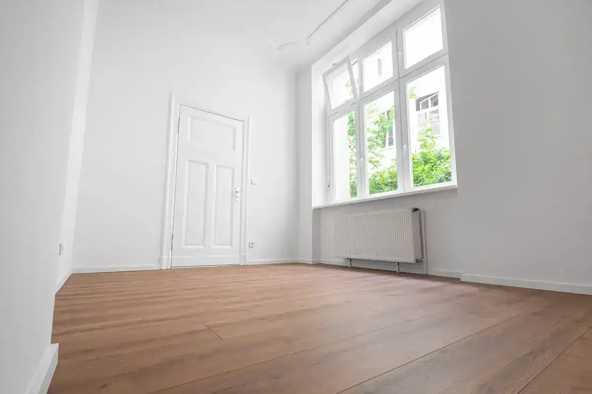 Puste, jasne pomieszczenie z białymi ścianami, drzwiami i oknem. Drewniana podłoga i grzejnik sugerują, że to gotowe do zamieszkania mieszkanie socjalne.