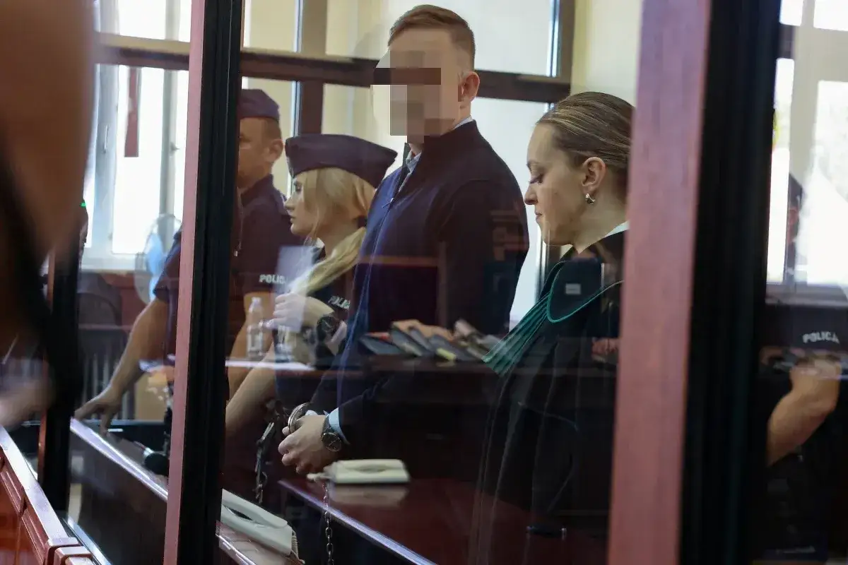 Osoba w kajdankach, prawniczka i policjanci w sali sądowej.