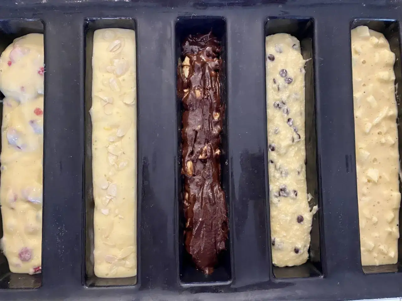 Cinq moules à cake longs prêts pour la cuisson : un aux fruits rouges, un aux amandes, un au chocolat, un aux pépites de chocolat et un aux pommes. Recette 5 cakes longs sucrés.