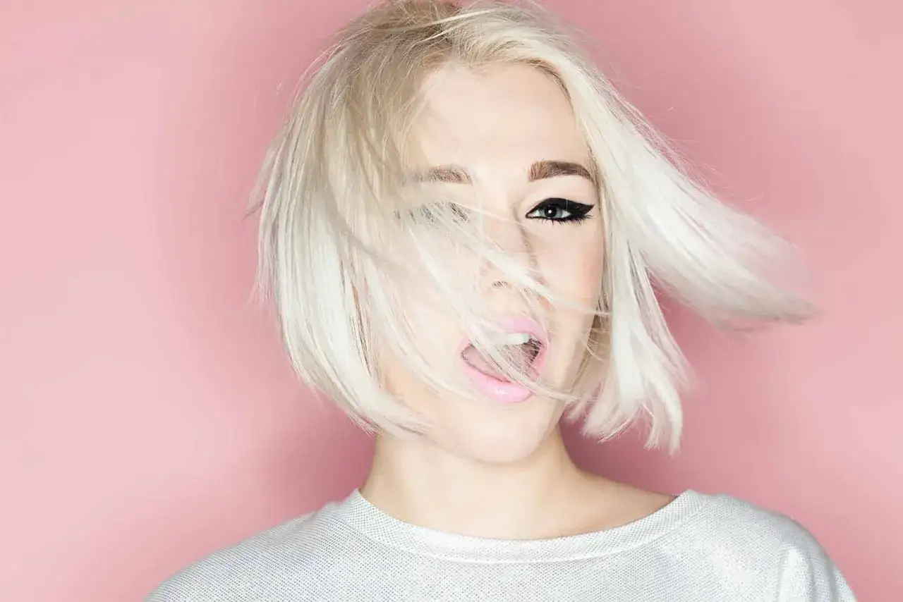 Mujer con cabello rubio platino y corte recto al hombro, con un ojo visible y labios rosados abiertos.