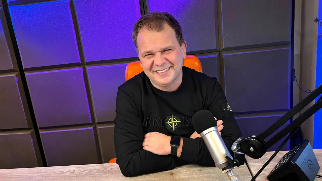 Uśmiechnięty mężczyzna w czarnej bluzie siedzi przy biurku z mikrofonem, na tle fioletowej ściany.