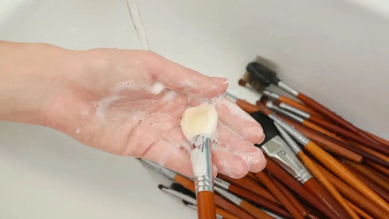 Mano con espuma jabonosa limpiando brochas de maquillaje en un lavabo.
