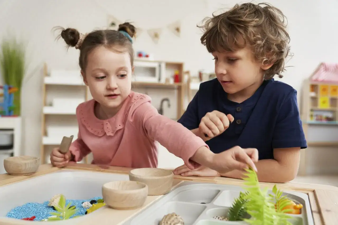 Dwoje dzieci bawi się w piaskownicy sensorycznej z niebieskim piaskiem i roślinami.