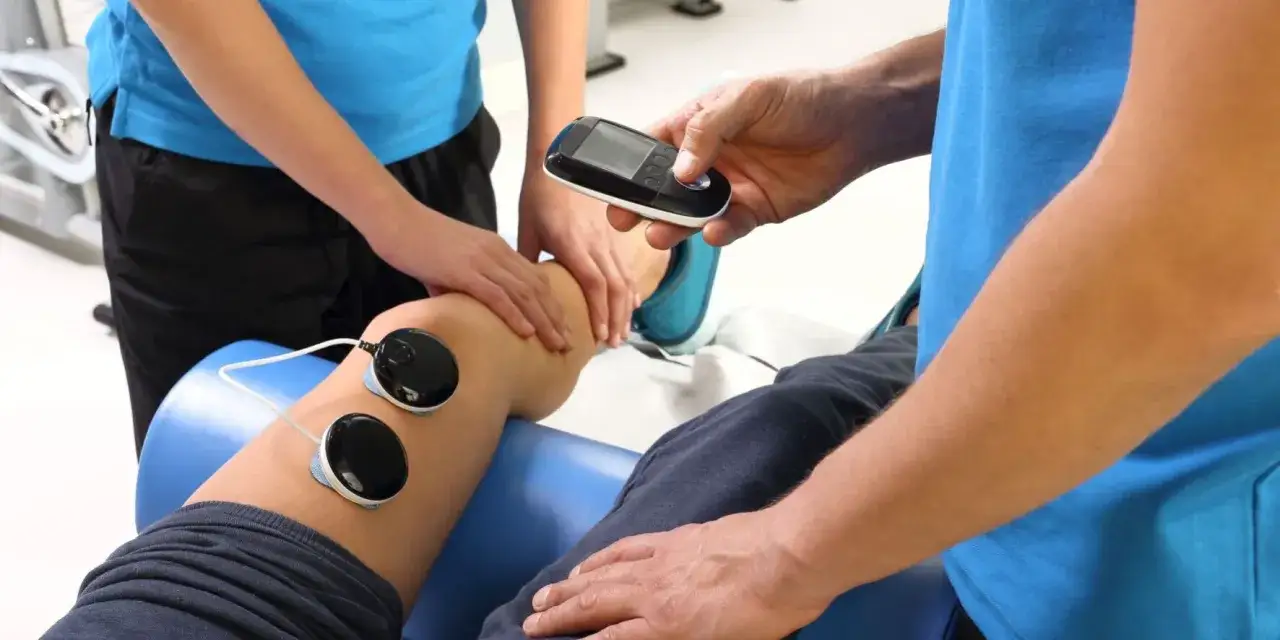 Rehabilitacja: fizjoterapeuta z urządzeniem do elektroterapii przy nodze pacjenta.