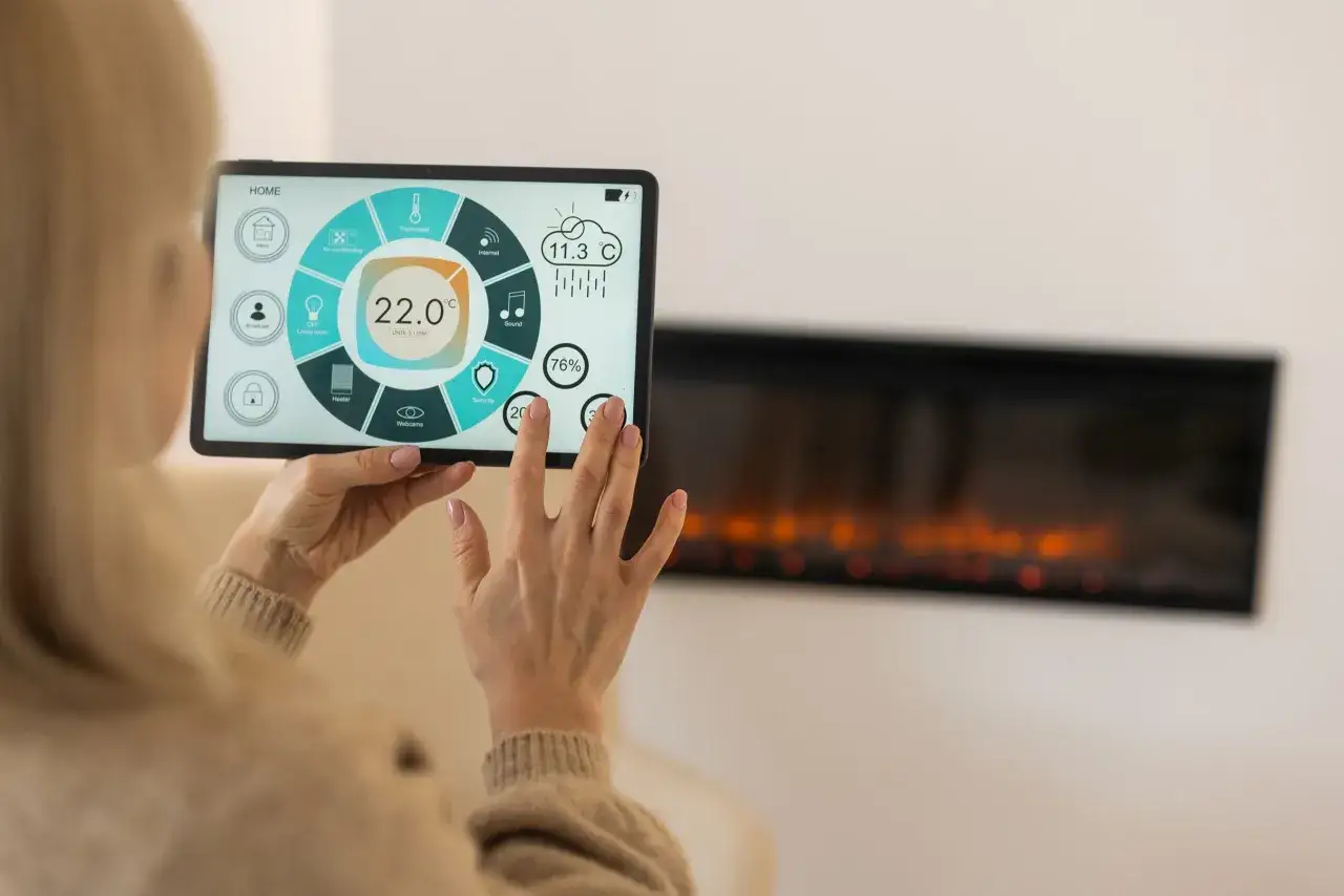Kobieta steruje inteligentnym domem przez tablet, kontrolując temperaturę i inne funkcje. Widoczny jest ekran z ikonami i wskaźnikami, a w tle kominek.