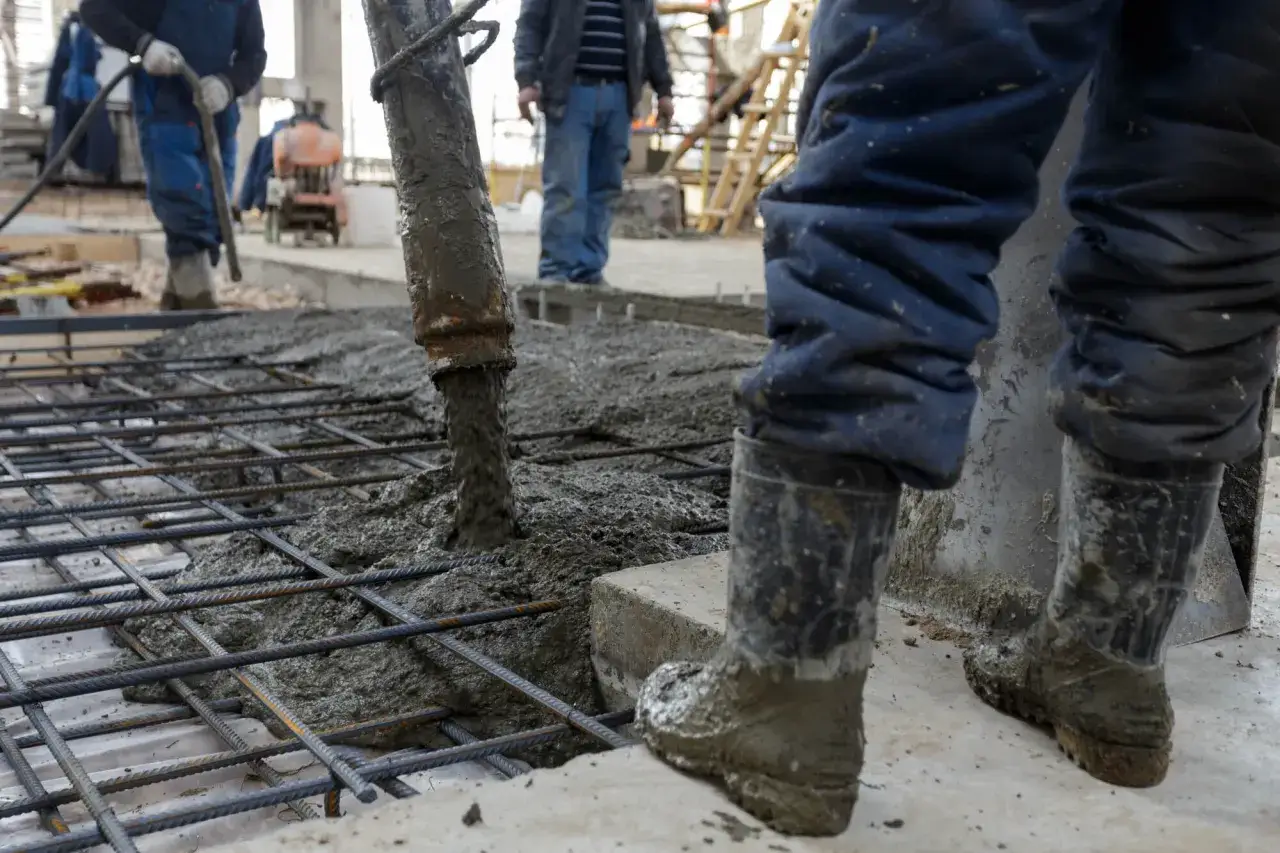 Betoniarka wylewa beton na zbrojenie, pracownicy w gumowcach nadzorują proces.