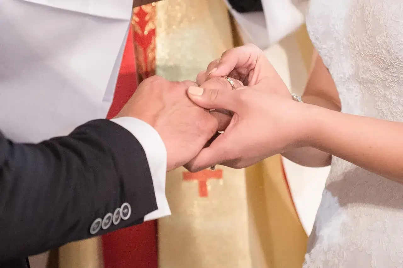 Dłoń panny młodej zakłada obrączkę na palec pana młodego. W tle widać księdza w szacie liturgicznej. To piękny ślub w innej parafii.