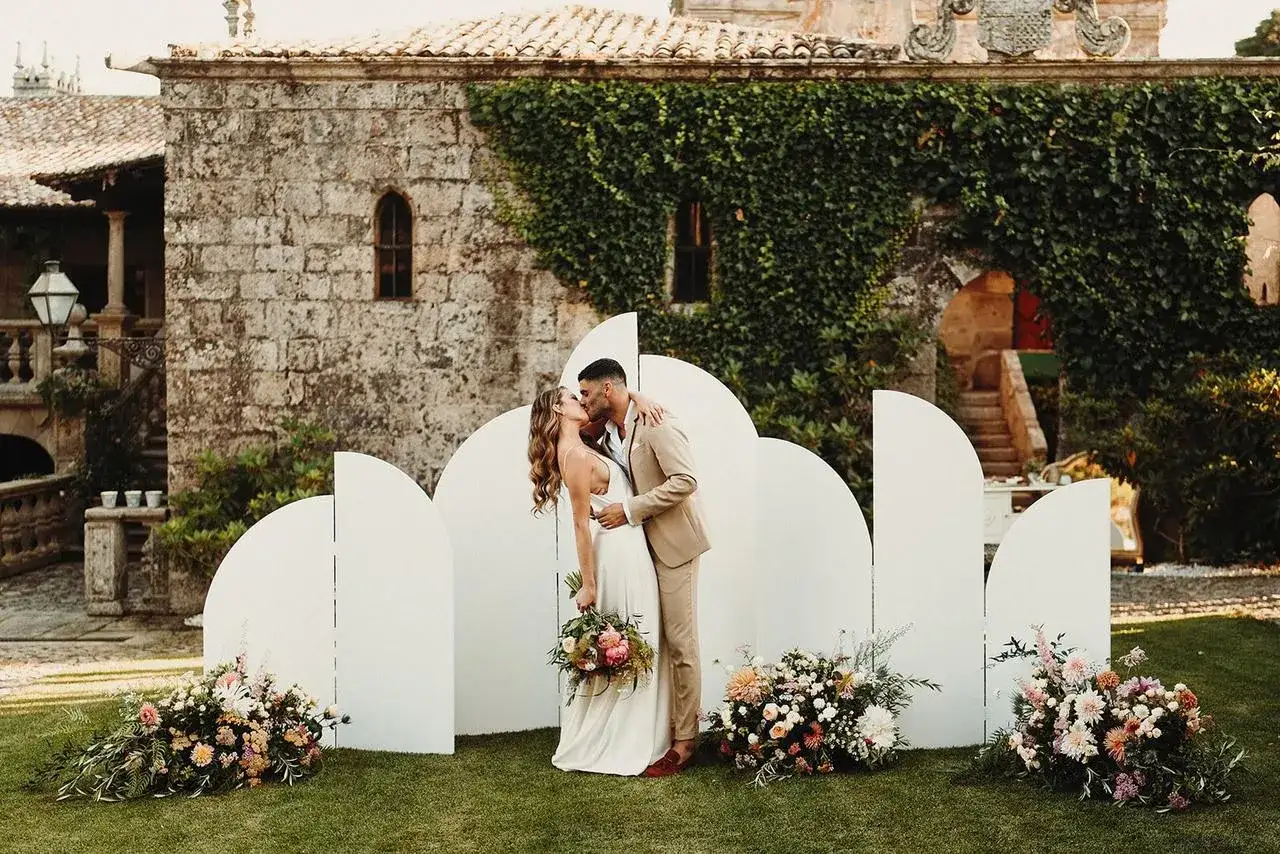 Novios besándose frente a un photocall moderno con arcos blancos y flores. ¡Ideas photocall boda únicas!