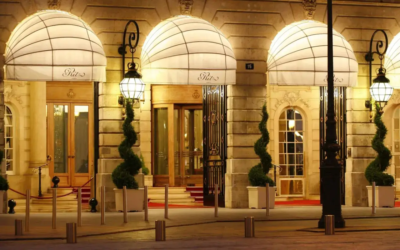 Eleganckie wejście do słynnego paryskiego hotelu Ritz, oświetlone nocą, z ozdobnymi markizami i spiralnymi drzewkami.