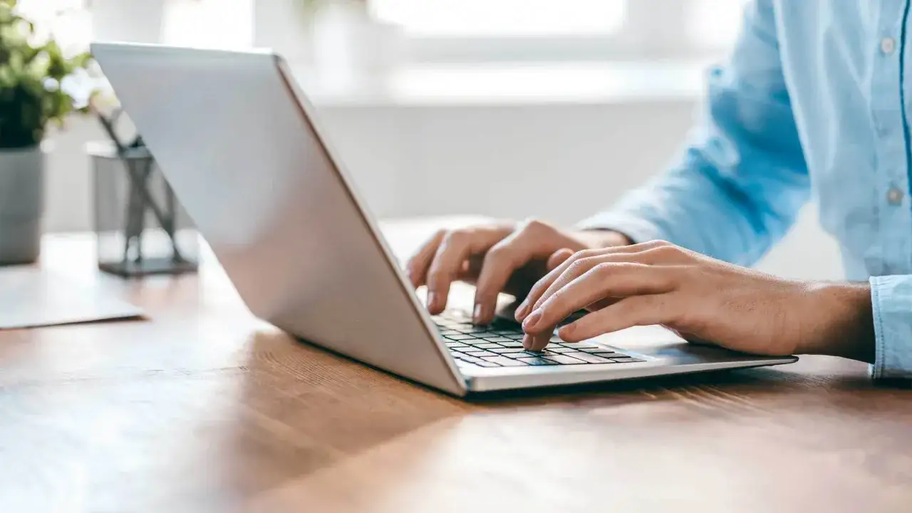 Person tippt auf einem silbernen Laptop auf einem Holztisch.