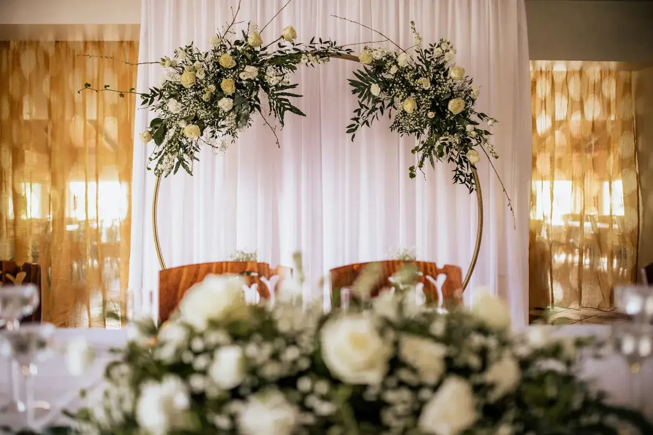 Elegancki ołtarz na ślub w plenerze, ozdobiony białymi kwiatami i zielenią na złotym okręgu.