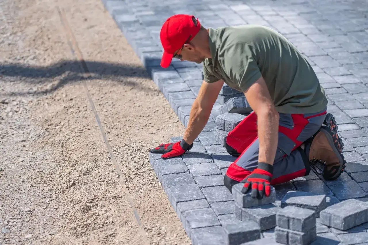 Pracownik w czerwonej czapce układa kostkę bauma na podsypce.