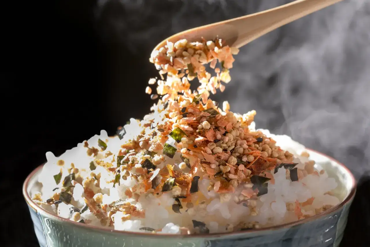 Łyżka sypie na parujący ryż kolorowy furikake. To japońska posypka, która dodaje smaku.