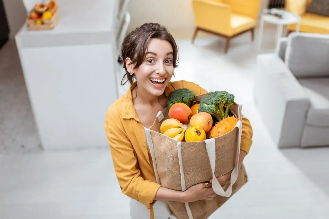 Uśmiechnięta kobieta z torbą pełną świeżych owoców i warzyw. Dobry wybór produktów to klucz do zdrowia i satysfakcji, unikaj skutków kupowania produktów z dołu rankingu opłacalność vs jakość.