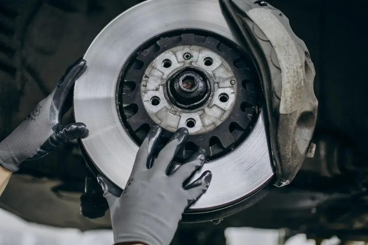 Mechanik w rękawiczkach wymienia zużyte tarcze hamulcowe. Widać nowy, lśniący element obok starego zacisku.
