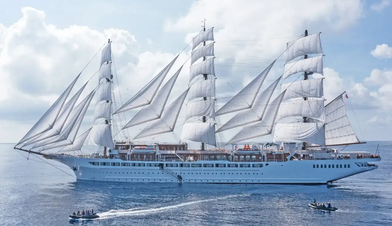 Ein majestätisches Segelschiff mit vollen weißen Segeln, das wie ein **sea cloud spirit** über das blaue Meer gleitet. Kleine Beiboote sind in der Nähe.