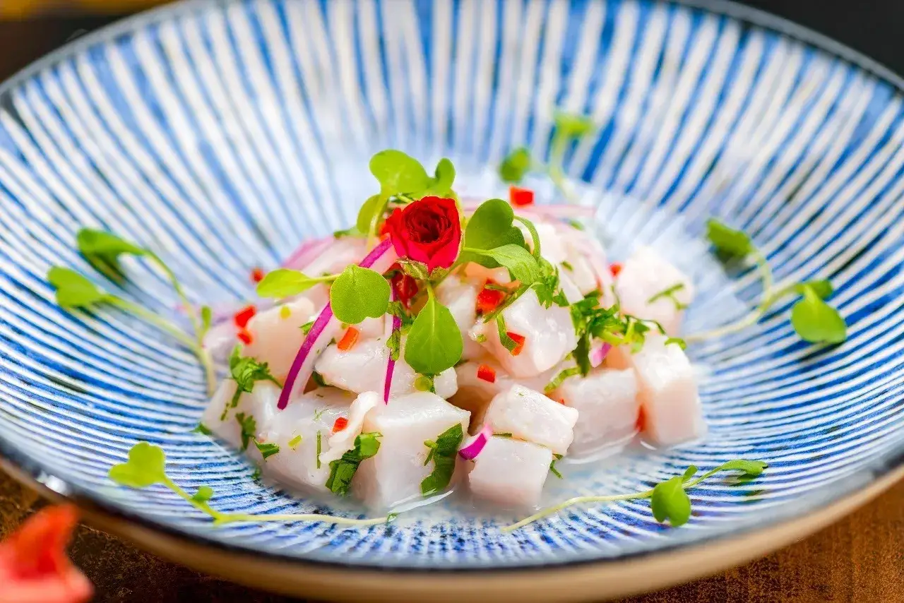 Pyszne ceviche z kawałków białej ryby, czerwonej cebuli, papryczki chili i ziół, udekorowane kiełkami i małą czerwoną różą, na niebiesko-białym talerzu.