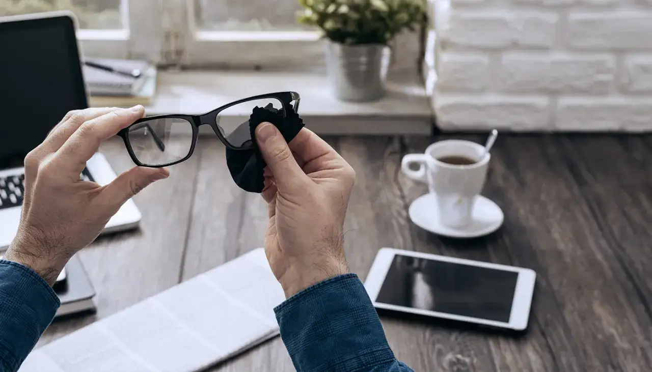 Mężczyzna czyści okulary z antyrefleksem czarną ściereczką. Na biurku leży laptop, tablet i filiżanka kawy.