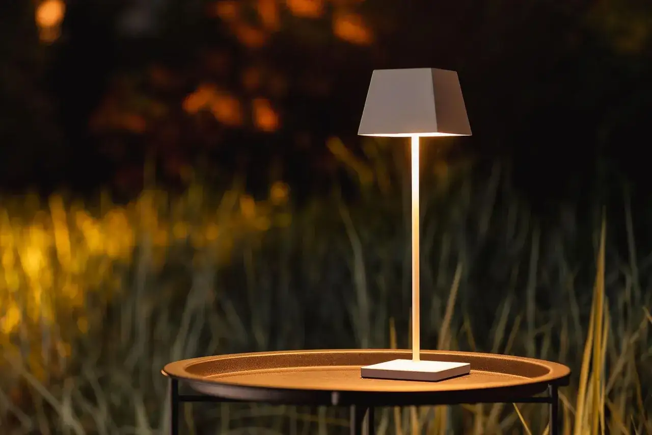 Nowoczesna lampa na balkon, stojąca na stoliku, oświetla wieczorny ogród.