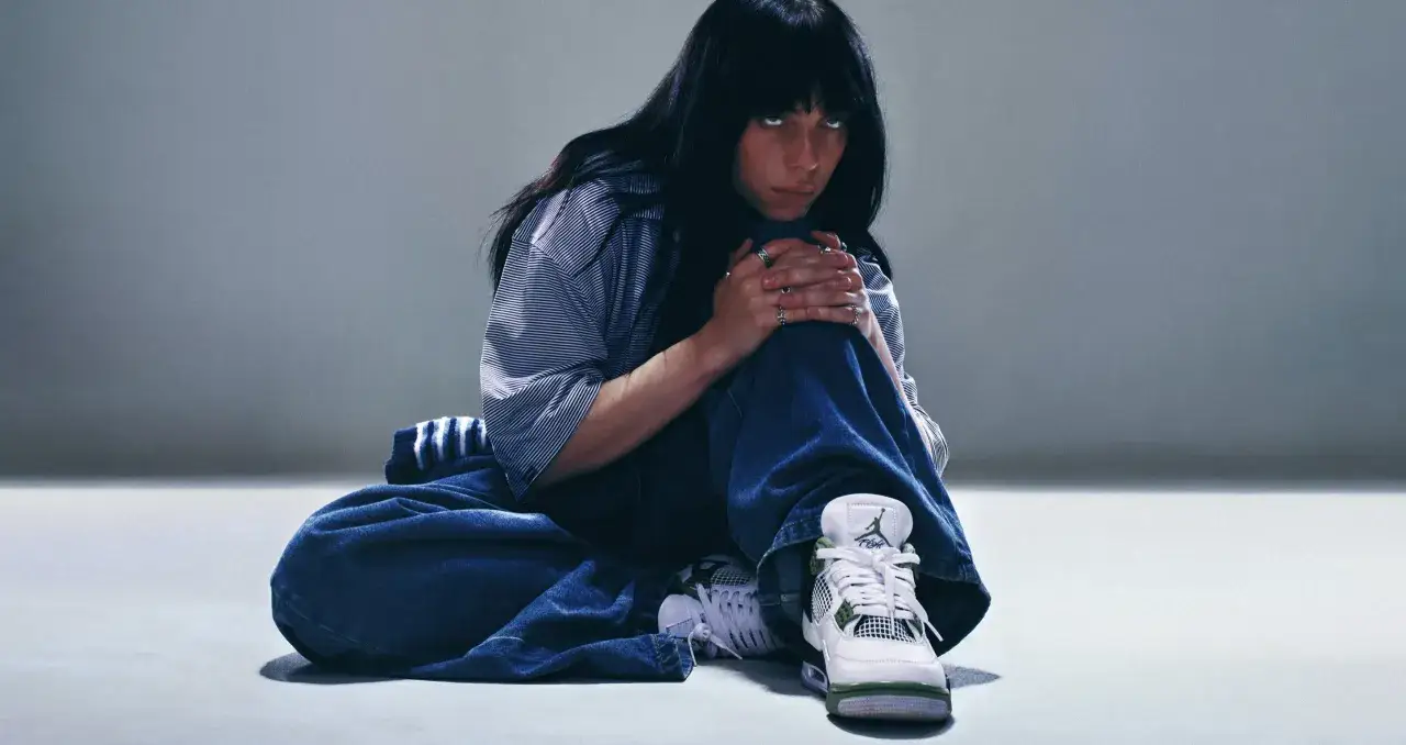 Billie Eilish w luźnych dżinsach i koszuli, zamyślona, w butach Jordan. Tekst "What was I made for?" pasuje do jej nastroju.