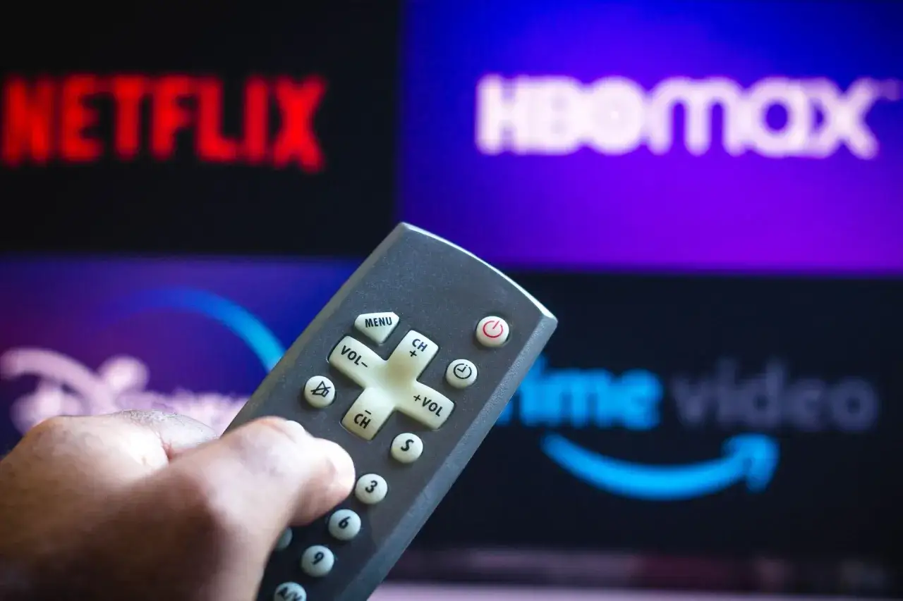 Dłoń trzymająca pilot do telewizora na tle ekranu z logo HBO Max, Netflix i Prime Video.