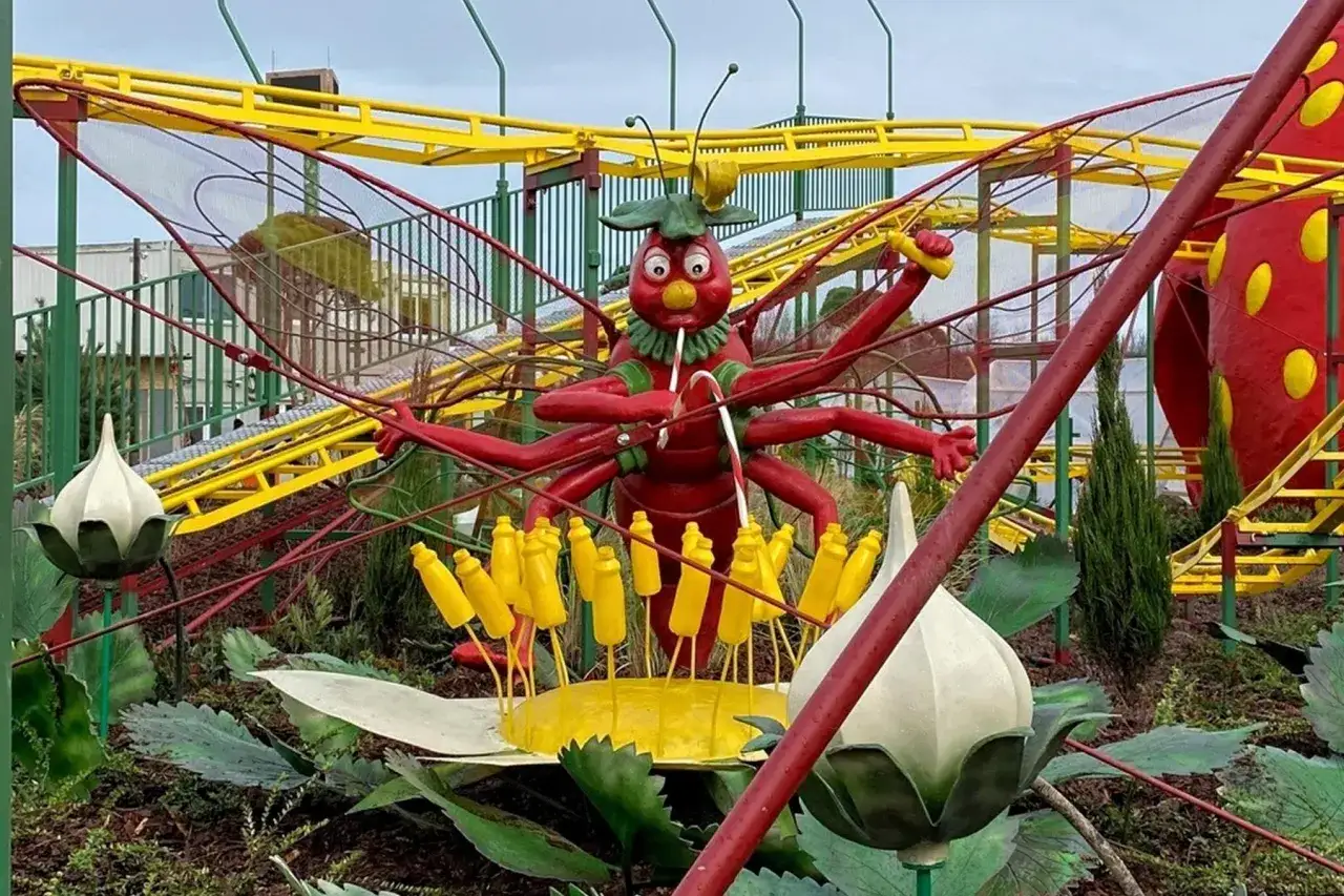 Eine rote Insektenfigur mit vielen Armen steht vor einer gelben Achterbahn.