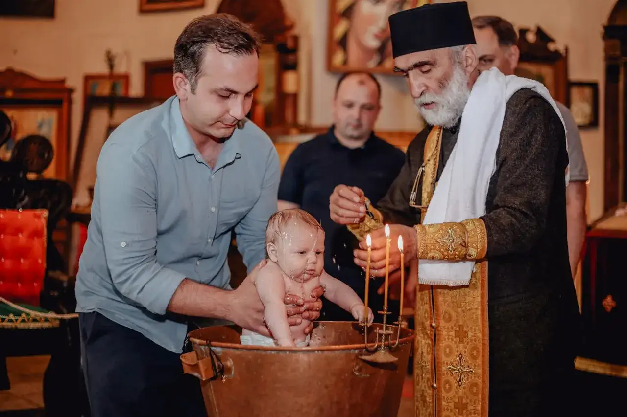 Chrzest dziecka w kościele. Ojciec trzyma niemowlę w miedzianej chrzcielnicy, a ksiądz polewa je wodą. To ważne chrzciny.