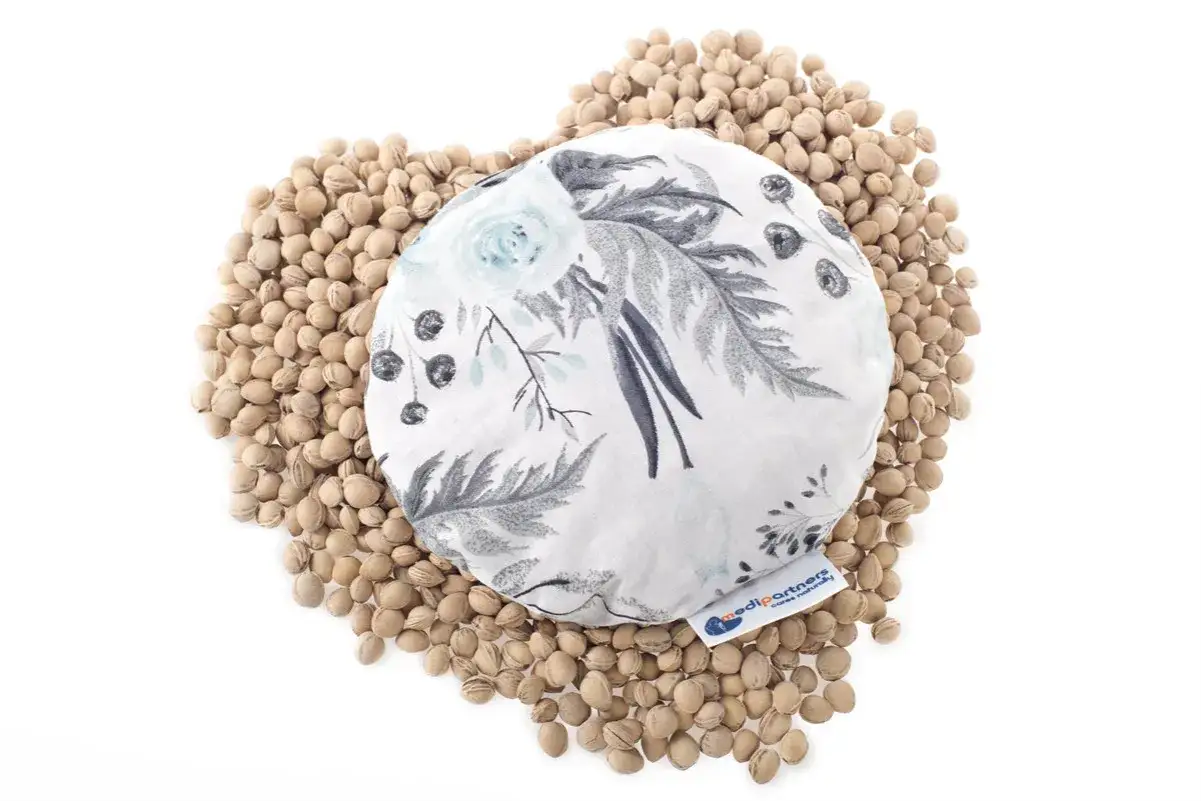 Okrągły termofor z pestek wiśni dla noworodka, ozdobiony delikatnym wzorem kwiatowym. Otoczony przez mnóstwo pestek wiśni.