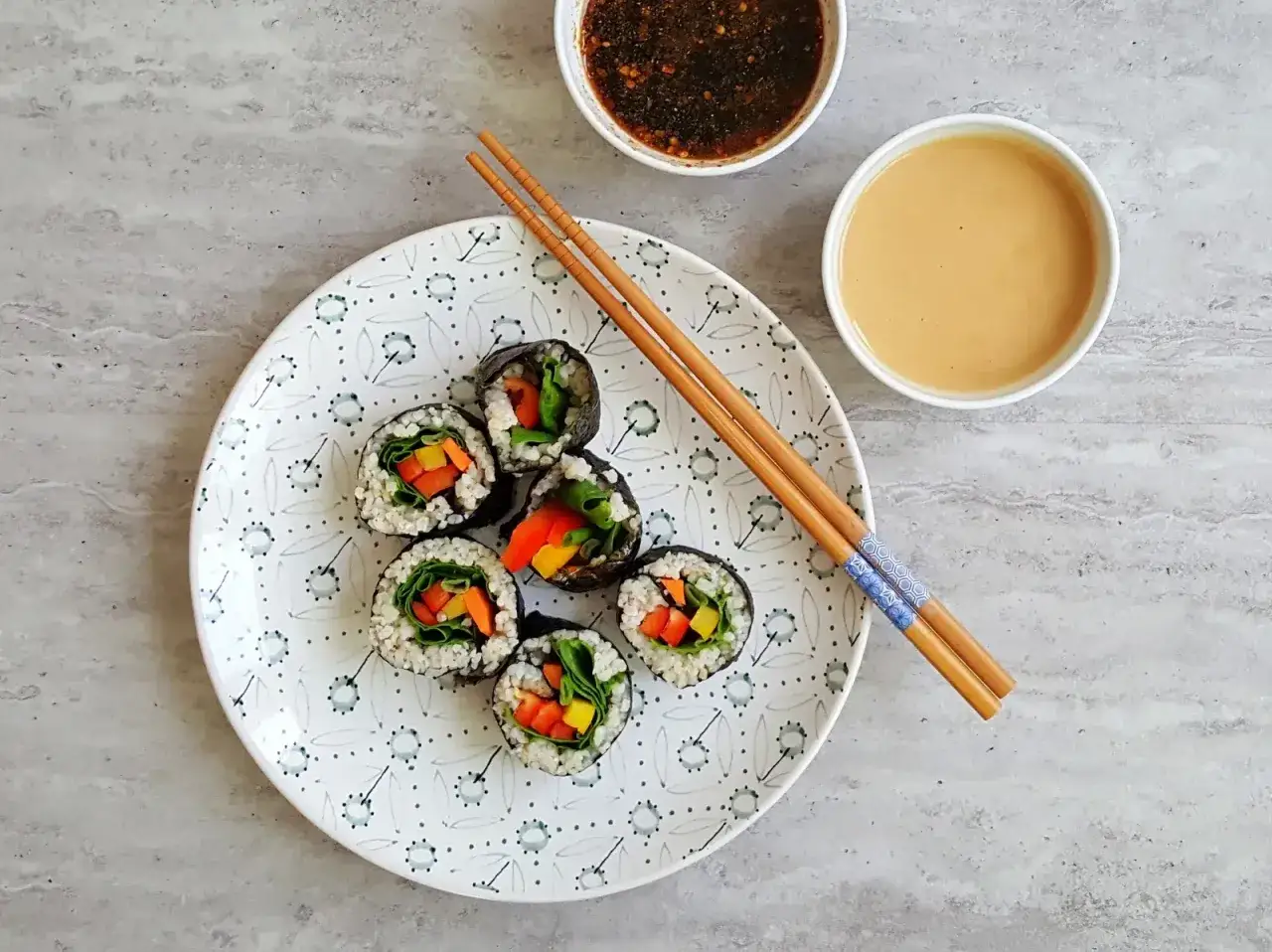 Sześć wegetariańskich rolek sushi z kolorowymi warzywami, przygotowanych z użyciem glonu do sushi, leży na talerzu obok sosów i pałeczek.